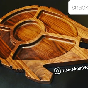 Puede incluir: Una bandeja de aperitivos de madera con forma de Halcón Milenario de Star Wars. La bandeja tiene cinco compartimentos para aperitivos y salsas. La bandeja está hecha de madera oscura y tiene un acabado liso y pulido.