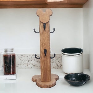 May include: Wooden mug rack shaped like Mickey Mouse with black metal hooks. The rack is standing on a white countertop.