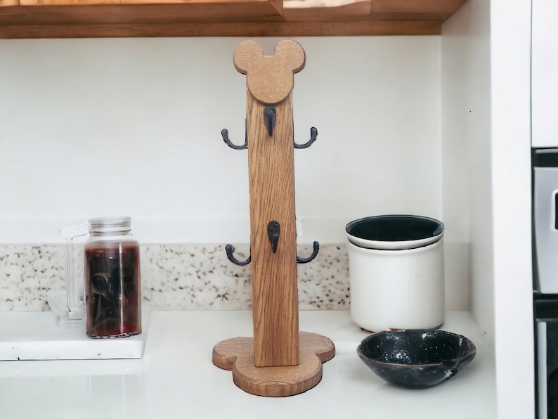 May include: Wooden mug rack shaped like Mickey Mouse with black metal hooks. The rack is standing on a white countertop.