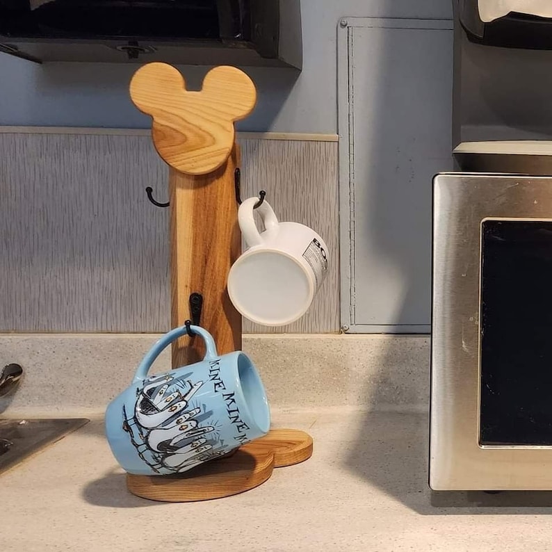 May include: Wooden mug rack shaped like Mickey Mouse head with two hooks on each side. The rack has a blue mug with a penguin design and a white mug hanging from it.