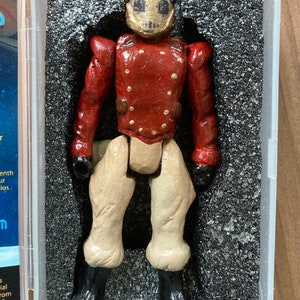 May include: A red and gold articulated action figure with a helmet and black boots. The figure is posed standing in a clear plastic display case with black foam padding.