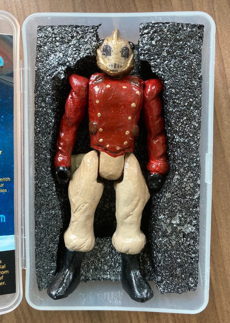 May include: A red and gold articulated action figure with a helmet and black boots. The figure is posed standing in a clear plastic display case with black foam padding.