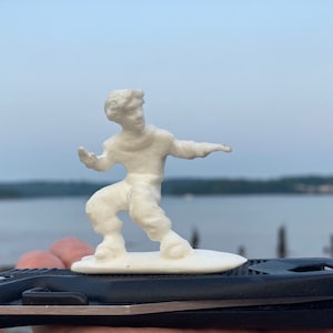 May include: A white 3D printed figurine of a person in a dynamic pose, standing on a small white platform. The figurine is posed as if surfing or skateboarding.