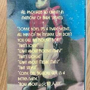 May include: A red and silver robot figure with a helmet and a large, round head. The figure is standing on a blue and white background. The text on the image reads: "ALL PROCEEDS TO CHANITY IN MEMORY OF DAVE STEVENS" and "SORRY, BOYS. IT'S A TRADE SECRET.... ALL PART OF THE MYSTERY. WHY DON'T YOU CALL HIM ROCKET BOY?" "THAT'S LOUSY!" "WHAT ABOUT HOCKET MAN?" "THAT'S WORSE!" "WHAT ABOUT MISSILE MAN?" "THAT STINKS!" "COME ON, BIGELOW, GIVE US A BETTER NAME." "HOW ABOUT 火箭人???"