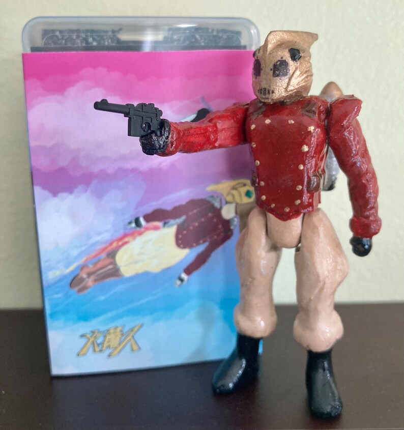 May include: A hand-painted action figure of a robot wearing a red and gold uniform, holding a black gun. The figure is standing in front of a pink and blue background with the Japanese word "炎" written in black.