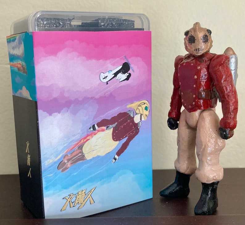 May include: A red and brown action figure with a jetpack and a helmet with a face mask. The figure is standing in front of a box with a pink and blue sky background and the text "火葬人" in black and gold.