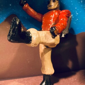 May include: A hand-painted figurine of a robot wearing a red and gold uniform with black boots. The robot is standing with one leg raised and its arm raised in the air.