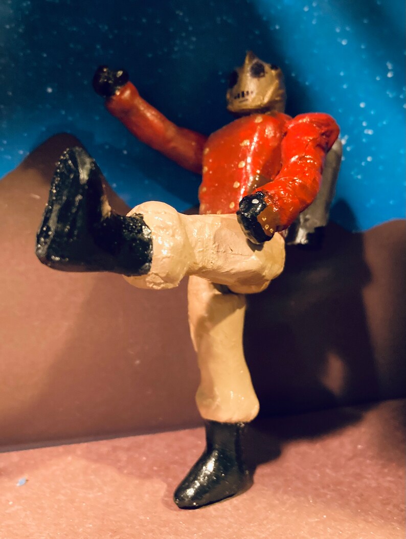 May include: A hand-painted figurine of a robot wearing a red and gold uniform with black boots. The robot is standing with one leg raised and its arm raised in the air.