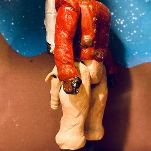 May include: A red and white painted action figure with a silver jetpack. The figure has a gold helmet with a skull design and is wearing beige pants and black boots.