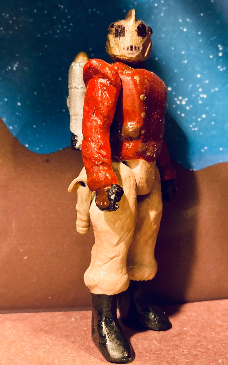 May include: A red and white painted action figure with a silver jetpack. The figure has a gold helmet with a skull design and is wearing beige pants and black boots.
