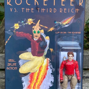 May include: A custom action figure of The Rocketeer, a superhero with a jetpack, in a red shirt and brown pants. The figure is in a plastic blister pack with a black and white background. The background image shows The Rocketeer flying through the air with flames coming from his jetpack. The text on the card reads "The Rocketeer vs. The Third Reich" and "Real Zipline Action!"