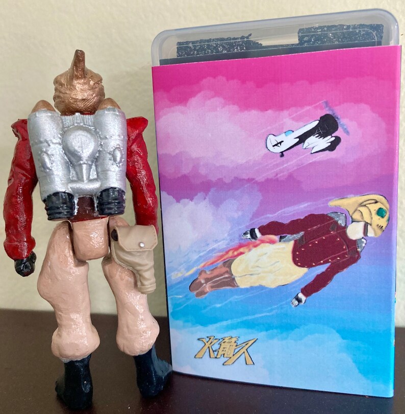 May include: A toy action figure with a silver jetpack and a red and brown outfit. The figure is standing in front of a pink and blue card with a cartoon image of a person flying with a jetpack and the text "火舞人".