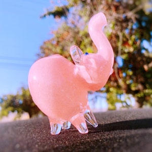 May include: A pink glass elephant figurine with a blue eye. The elephant has a raised trunk and is standing on a dark surface.