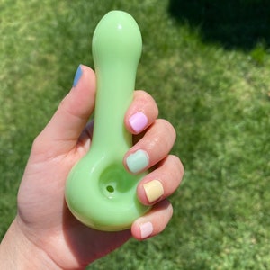 May include: A light green glass pipe with a rounded bowl and a small hole for smoking. The pipe is being held in a hand with pastel-colored nail polish.