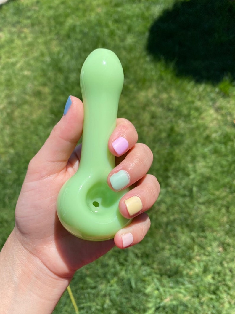May include: A light green glass pipe with a rounded bowl and a small hole for smoking. The pipe is being held in a hand with pastel-colored nail polish.