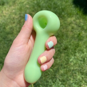 May include: A light green glass pipe with a rounded bowl and a long, slender stem. The pipe is held in a hand with pastel-colored nail polish.