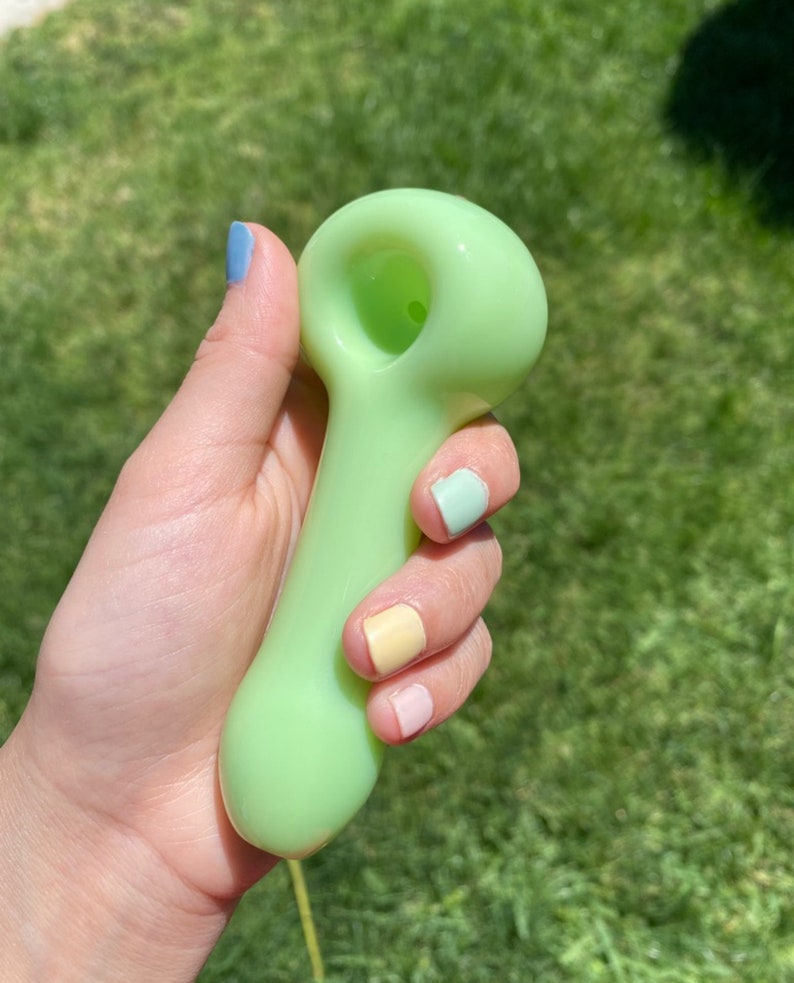 May include: A light green glass pipe with a rounded bowl and a long, slender stem. The pipe is held in a hand with pastel-colored nail polish.