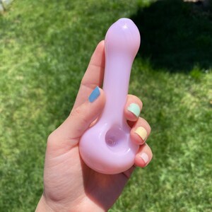 May include: A light purple glass pipe with a rounded bowl and a long, slender stem. The pipe is held in a hand with colorful nail polish.