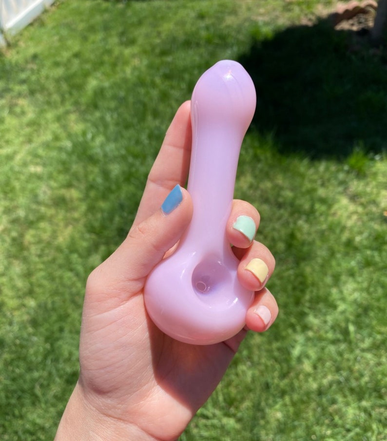 May include: A light purple glass pipe with a rounded bowl and a long, slender stem. The pipe is held in a hand with colorful nail polish.