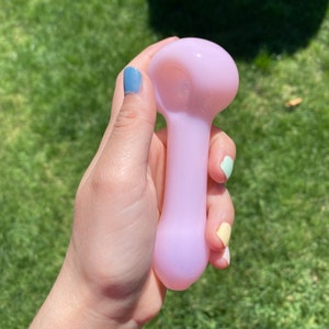May include: A light pink glass pipe with a rounded bowl and a straight stem. The pipe is being held in a hand with painted fingernails.