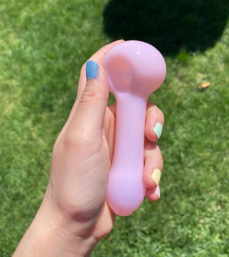 May include: A light pink glass pipe with a rounded bowl and a straight stem. The pipe is being held in a hand with painted fingernails.