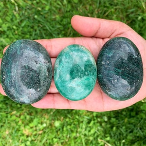 May include: Three smooth, green, polished stones, also known as worry stones, in various shades of green, are held in a hand.