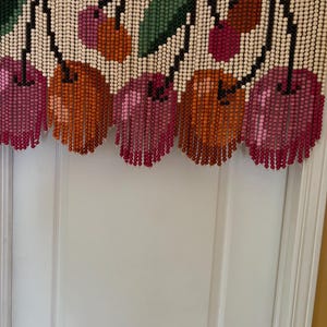 May include: A beaded cherry door curtain. The curtain features a white background with a design of cherries and green leaves. The cherries are in shades of pink, orange, and red. The curtain hangs in front of a white door.