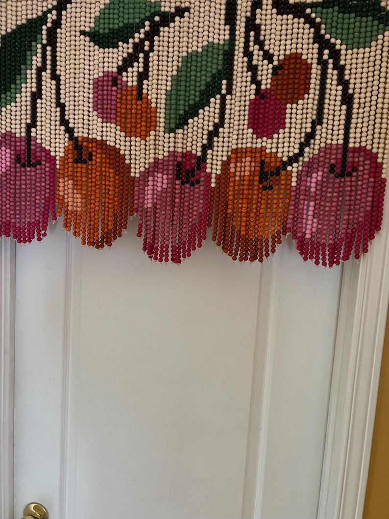 May include: A beaded cherry door curtain. The curtain features a white background with a design of cherries and green leaves. The cherries are in shades of pink, orange, and red. The curtain hangs in front of a white door.