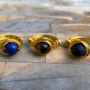 May include: Three gold wire wrapped rings featuring different coloured stones. The first ring has a blue lapis lazuli stone, the second ring has a brown tiger's eye stone, and the third ring has a black lava stone.