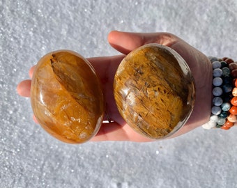 Puede incluir: Dos piedras de ojo de tigre pulidas, una con un tono dorado y la otra con un color más marrón y dorado, se muestran en una mano.