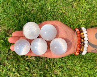 Op de afbeelding: Vijf gladde, witte seleniet bollen, elk met een licht verschillend patroon van strepen, worden in een hand gehouden. De bollen hebben een diameter van ongeveer 5 cm.