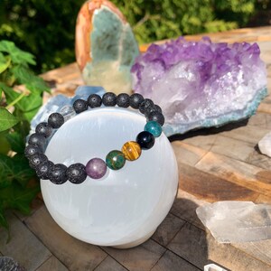 Op de afbeelding: Een zwarte lava armband met gekleurde edelsteen kralen, waaronder amethyst, tijgeroog en groene aventurijn. De armband wordt getoond op een witte seleniet bol.