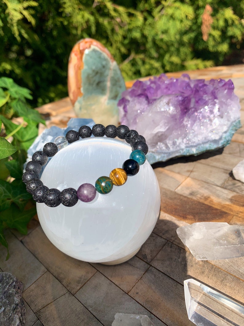 Op de afbeelding: Een zwarte lava armband met gekleurde edelsteen kralen, waaronder amethyst, tijgeroog en groene aventurijn. De armband wordt getoond op een witte seleniet bol.