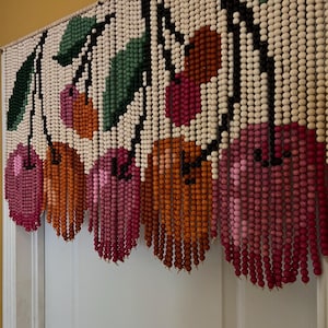 May include: A beaded curtain featuring a cherry design. The curtain has white beads as a background, with black stems and green leaves. The cherries are in shades of pink and orange, creating a colorful and decorative accent.