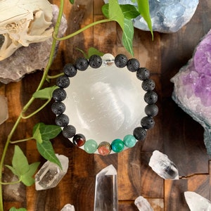 May include: A black lava rock bracelet with red tiger's eye, green aventurine, pink rose quartz, and green turquoise beads. The bracelet is on a white surface with crystals and greenery.
