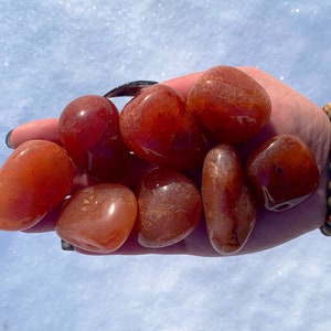 May include: A collection of eight carnelian tumbled stones in various sizes and shades of orange and red. The stones are smooth and polished, with a natural, earthy look.