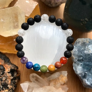 Könnte beinhalten: Ein Chakra-Armband mit schwarzen Lava-Perlen und klaren Quarz-Perlen. Das Armband enthält auch sieben Chakra-Steine in den Farben des Regenbogens.