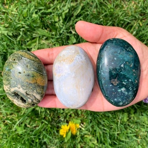 May include: Three smooth, polished, oval-shaped stones. The stones are green, white, and blue with brown and yellow accents. The stones are arranged in a row on a hand.