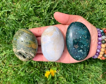 May include: Three smooth, polished, oval-shaped stones. The stones are green, white, and blue with brown and yellow accents. The stones are arranged in a row on a hand.