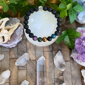 Op de afbeelding: Een zwarte lava armband met amethist, groene aventurijn, tijgeroog en blauwe apatiet kralen. De armband ligt op een wit oppervlak met heldere kwartskristallen.