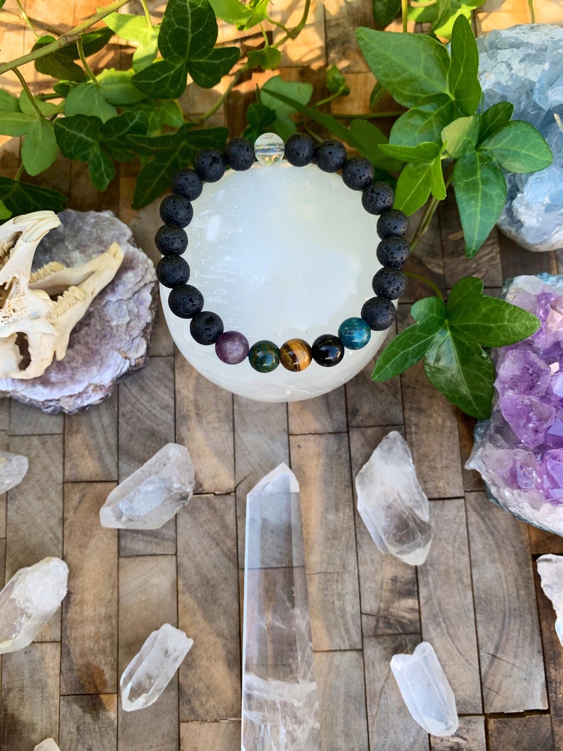 Op de afbeelding: Een zwarte lava armband met amethist, groene aventurijn, tijgeroog en blauwe apatiet kralen. De armband ligt op een wit oppervlak met heldere kwartskristallen.
