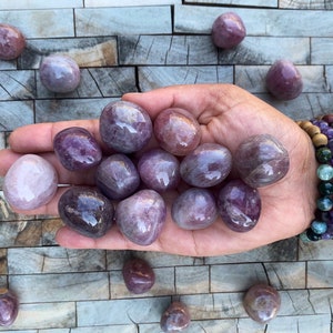 May include: A selection of polished amethyst stones in shades of purple and lavender. The smooth, oval-shaped stones have a translucent appearance. Some are held in a hand, others are scattered on a textured surface. The stones vary in size.