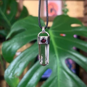 May include: A clear quartz crystal pendant with a silver setting and a red garnet accent. The pendant is on a black cord.
