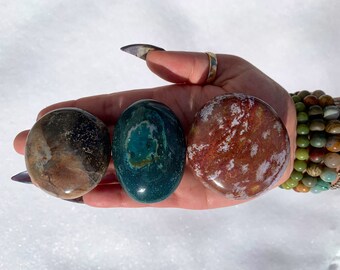 May include: Four smooth, polished stones in various colours and patterns. The stones are brown, green, and red with white and gold accents. The stones are arranged in a row on a hand.