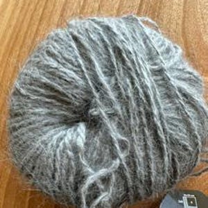 May include: A ball of gray yarn, tightly wound, with a soft, textured appearance. The yarn is a muted gray color, and the fibers appear to be slightly fuzzy. A small tag is visible at the bottom right.