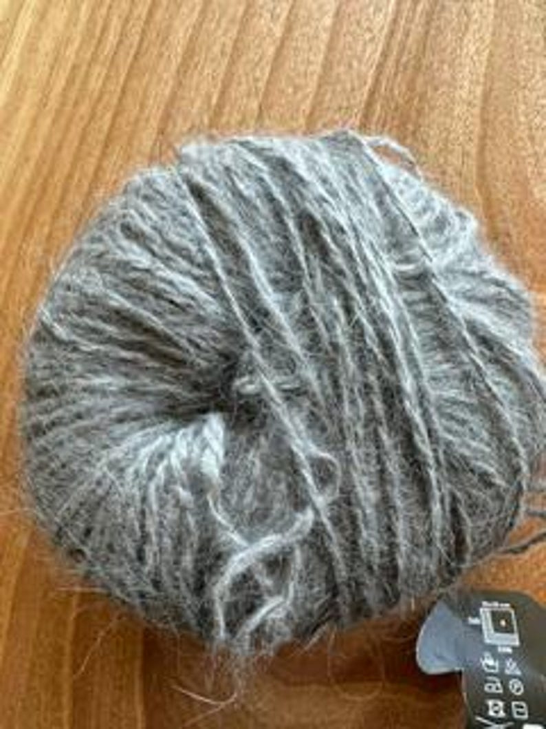May include: A ball of gray yarn, tightly wound, with a soft, textured appearance. The yarn is a muted gray color, and the fibers appear to be slightly fuzzy. A small tag is visible at the bottom right.