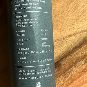 May include: A skein of yarn in a light brown color with a green and orange stripe. The yarn is labeled "Poilen" and has a dye lot number of 1419. The yarn is made of 48% recycled silk, 36% fine merino, and 16% cashmere. The yarn is light fingering weight and has a yardage of 224 yards / 205 meters. The gauge is 25 stitches = 4 inches on US size 3 needles. The yarn is made in Japan and is hand washable.