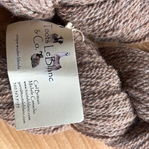 Op de afbeelding: Een bol bruin garen met een label waarop "Toots Le Blanc & Co." staat en contactgegevens voor Carl Brittain en Michele Camacho.