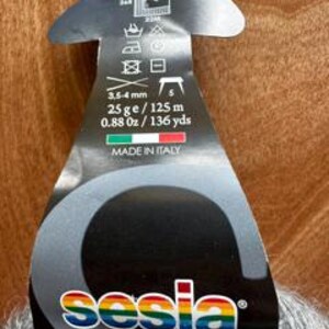 May include: A close-up of a yarn label with the brand name "sesia" in rainbow colors. The label is black and gray, with text indicating the yarn is 0.88 oz / 136 yds, 3.5-4 mm, and made in Italy. The yarn is gray.