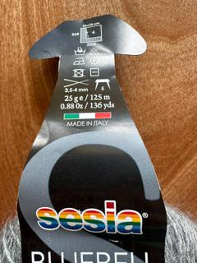 May include: A close-up of a yarn label with the brand name "sesia" in rainbow colors. The label is black and gray, with text indicating the yarn is 0.88 oz / 136 yds, 3.5-4 mm, and made in Italy. The yarn is gray.